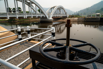 Black metal cogwheel controlling floodgate with texts in Thai language meaning "the property of", and the view of white concrete railroad bridge at the back. 