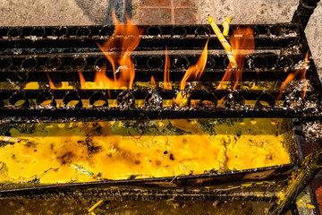 Yellow candles burning on black metal holder with orange flame and yellow candle tears underneath during daytime in a Buddhist temple. 