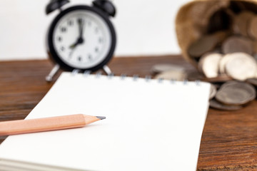 Blank notepad, alarm clock, coins and pencil laying on it on office wooden table