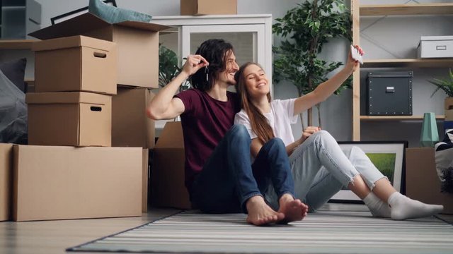 Girl and guy are taking selfie with keys in new house using modern smartphone camera posing and kissing enjoying new accommodation. Youth and photos concept.