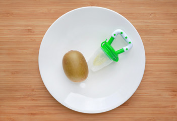 Baby's nibbler and kiwi on white plate against wooden background. Organic baby food concept.
