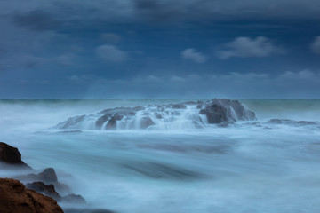 Bei Sturm und dunklem Wolkenhimmel überspülen Wellen einen Felsen , fotografiert in Langzeitbelichtung