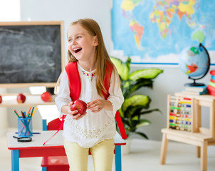 Little laughing blond girl holding an apple in the school classroom