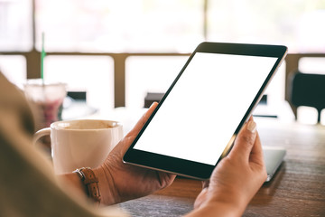 Mockup image of hands holding black tablet pc with blank white screen with coffee cup on wooden table