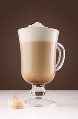 Cappuccino coffee with foam and cookies in transparent glass with handle on dark brown wall and white wood board.