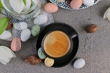 A cup of aromatic coffee with froth over a gray background, white tulips and chocolates for Easter. Copy space, flat lay.