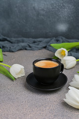 A cup of aromatic coffee with froth over a gray background and white tulips. copy space.