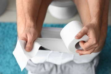 Man with roll of paper sitting on toilet bowl at home, closeup
