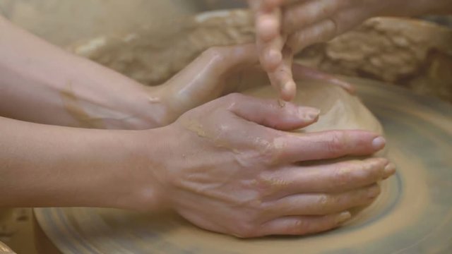 Two intertwined hands form a pot of clay on a potter's wheel. A male hand helps a woman's hands shape a clay shape.