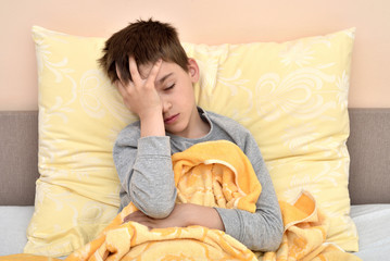 Ill cute young boy with closed eyes sitting in bed holding his head