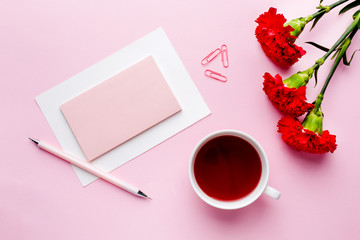 Red-pink objects. Cup of tea, carnation flowers Notepad for text on pastel pink background. Copy space. Top view Flat lay