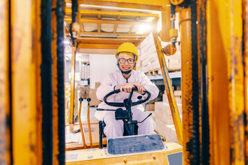 Fototapeta na wymiar Young beautiful Caucasian female woman in white uniform and with protective helmet on head driving forklift in warehouse.