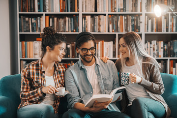 Three multicultural students sitting on sofa in library and talking about book. Man holding book...
