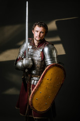Fototapeta na wymiar Medieval knight in armor holding sword and shield on the dark background