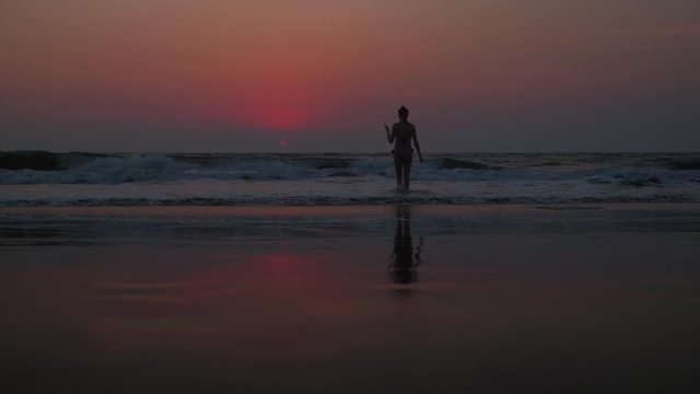 Silhouette of a beautiful young women in a bikini walking into the waters of the Arabian sea towards the beautiful setting sun with her mobile phone camera in hand for taking better photos and videos 