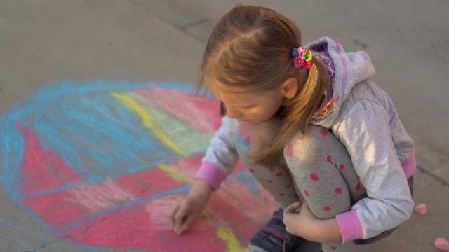 Happy baby fair-haired beautiful girl draws on the pavement with chalks
