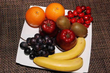 Fresh Oranges, apples, grapes, kiwis, cherries, bananas on the white plate on the brown table