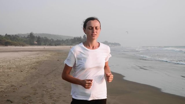 Woman with bandage on injured knee is running on the sandy ocean beach, stopping in pain and holding her knee, front view. Slow motion.
