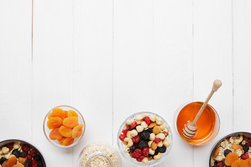 top view of bowls with cereal, dried apricots and berries, honey and nuts on white table