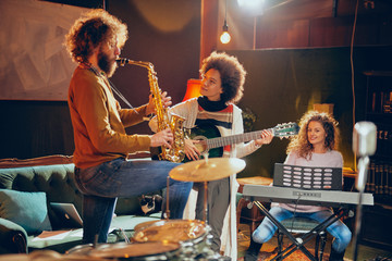 Mixed race woman playing acoustic guitar while man playing saxophone. Home studio interior. In...