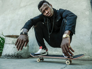 young attractive and serious black African American man squatting on skate board at grunge street...