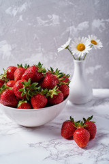 Red ripe strawberries in white ceramic bowl on linen table napkin and chamomile flowers in vase on grey plaster background, copy space. Healthy food concept, still life