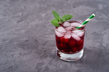 Fresh cherry lemonade with ice, mint and green paper straw in sparkling glass on grey plaster background, copy space. Cold summer drink. Berry cocktail