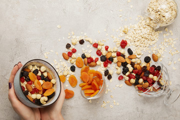 partial view of woman holding bowl with muesli, dried apricots and berries, nuts on textured grey...