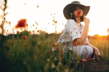 Stylish girl in linen dress smelling poppy flower in meadow in sunset light with flowers in rustic...