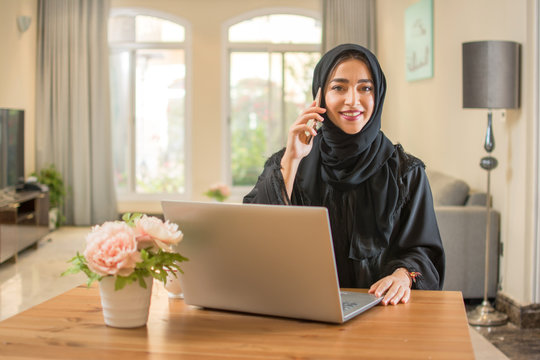 Beautiful young Arab woman wearing abaya talking on mobile phone and using laptop at home