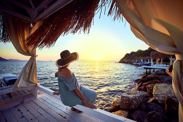 Woman in hat relaxing by the sea in a luxurious beachfront hotel resort at sunset enjoying perfect...