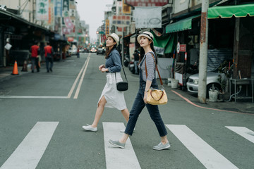 two happy female friends travelers with bags crossing street together outdoor sunny day in china town. japanese lady travel in chinese city walking on zebra cross in urban. girl wear hats look sky.