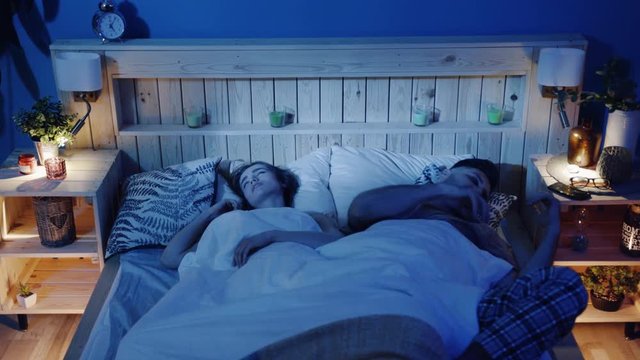 Young couple in bed, snoring woman, man cannot sleep, covering ears with pillow for snore noise, sleeping in bedroom at home. Man is trying to fall asleep while woman is snoring. Health issues problem