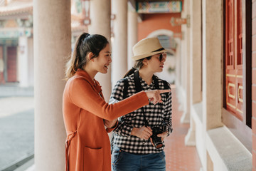 foreign female visitors looking inside room of historic building. smiling smart girl points finger explains to friend in sunglasses. two asian women tourists sightseeing chinese temple beijing china