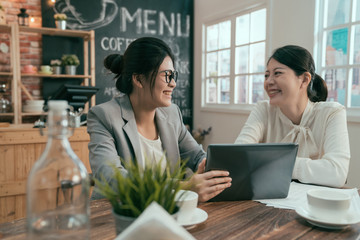 two beautiful young coworkers sitting in modern coffee shop interior and talking with happy smiles. Successful attractive businesswomen working cheerful on project in cafe by window on tablet.