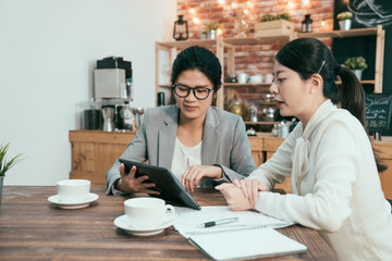 office ladies business cooperation meeting teamwork concept. Two businesswomen talk chat sitting at table in cafe shop. Girl in suit shows colleague information on tablet screen working in coffee bar