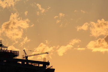Silhueta de guindaste e edifício em construção no pôr-do-sol com o céu repleto de nuvens na cor laranja