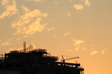 Silhueta de guindaste e edifício em construção no pôr-do-sol com o céu repleto de nuvens na cor laranja