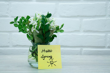A vase with snowdrops is standing on a wooden table, against a white loft with a pattern of the sun on a sticker and the text hello spring. Snowdrops in a vase on a wall background.