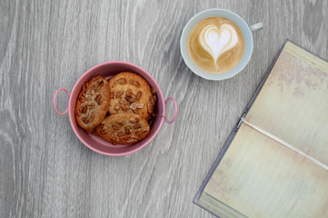A cup of coffee with a froth in the shape of a heart with cookies and an open diary on a wooden gray table. Breakfast.