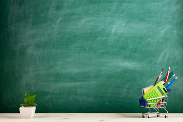 Education concept - school supplies in a shopping cart on the desk in the auditorium, blackboard background © Sensay