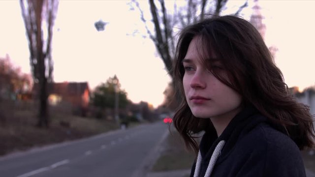 A girl is standing by the side of the road, she looks at the camera, then away.