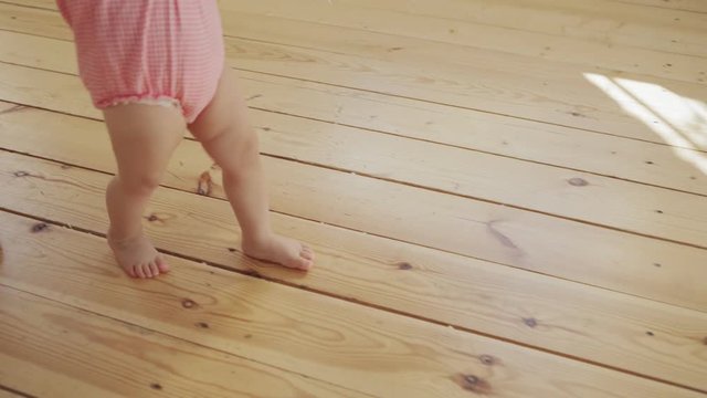 Tracking shot of unrecognizable caring mother supporting her adorable baby daughter learning to walk and making her first steps on hardwood floor at home