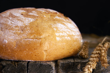 Round white bread and wheat ears on the old wooden table in the dark
