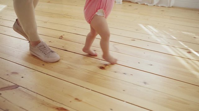 Tracking right shot of mother supporting her barefoot baby daughter in pink bodysuit learning to walk on hardwood floor at home