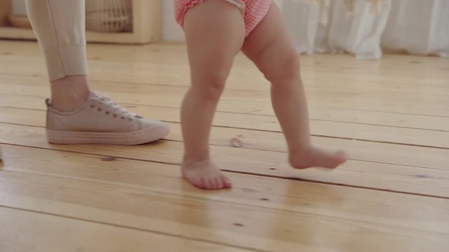 Tracking close-up shot of mother helping her barefoot baby daughter in pink bodysuit to make first steps at home