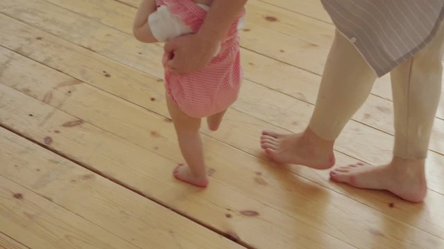 High angle view of cute baby girl in pink bodysuit making her first steps helped by caring mom at home