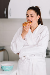 Woman having a breakfast in a robe at home. Girl eating a bagel at home.