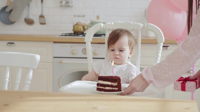 Loving mother giving baby daughter sitting in highchair a piece of birthday cake with one burning candle on her first birthday celebration