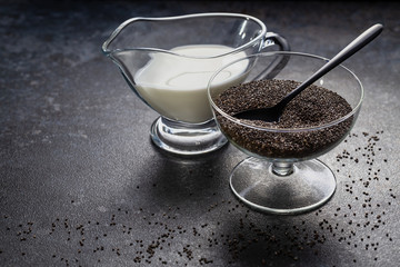 Chia seeds in a glass vase and coconut milk against a dark stone surface with scattered grains.